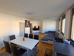 Un espace de vie moderne comprenant un coin repas, un ensemble de canapés, un téléviseur à écran plat et une fenêtre avec des rideaux.