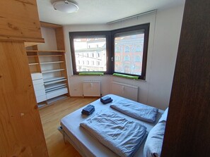 Een slaapkamer met een bed, een houten kledingkast en een raam met uitzicht op gebouwen.
