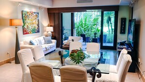Een moderne woonkamer met een glazen salontafel, witte meubels, een groot schilderij en uitzicht op groen.