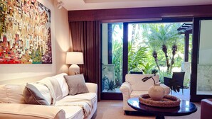 A modern hotel room with a white sofa, a side table with a vase, and a large window with a view of tropical plants.