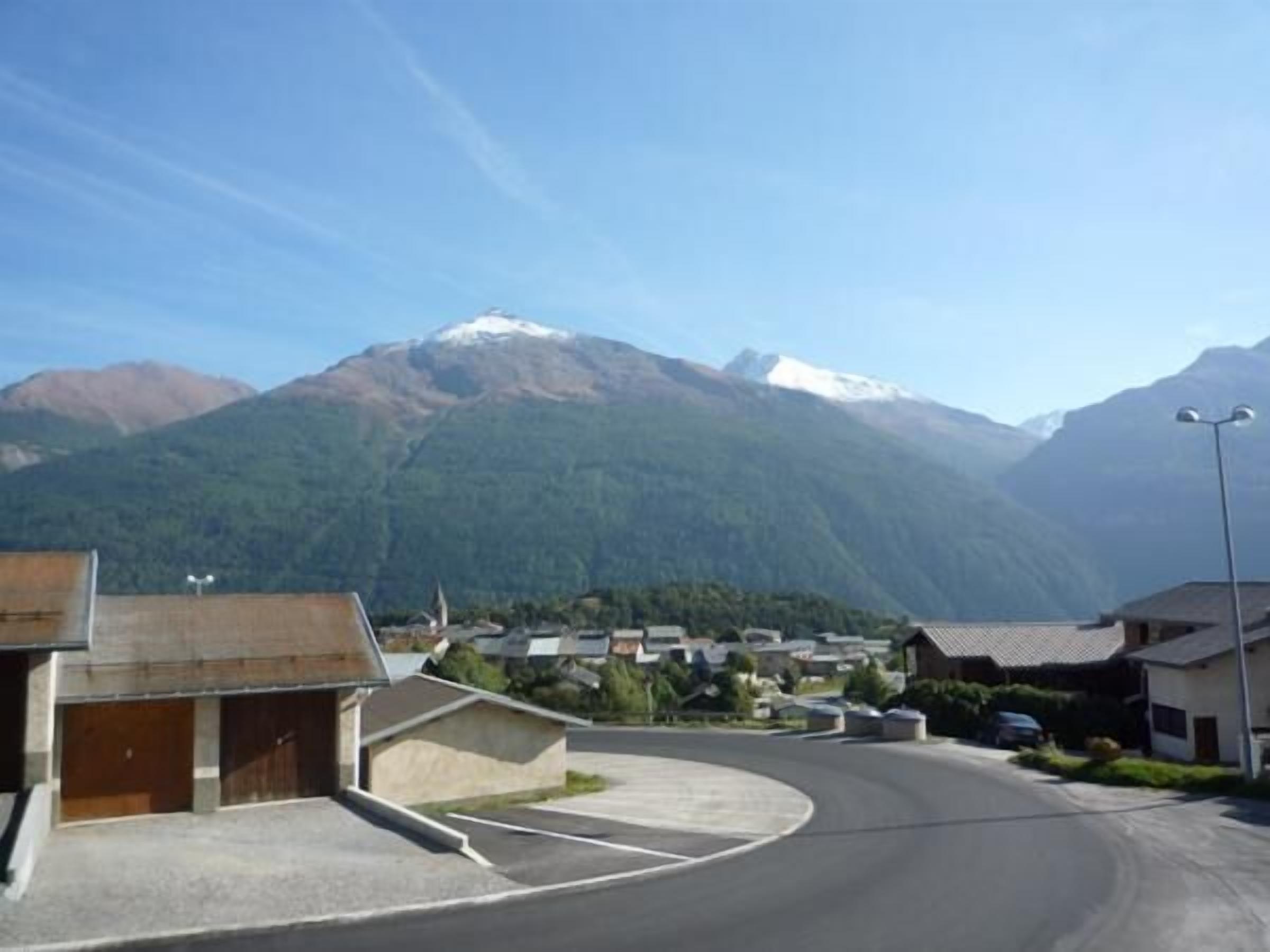 A residential area with houses, a curved road, and a mountain with snow-capped peaks in the background.