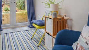 Une pièce avec un canapé bleu, un fauteuil jaune, un meuble en bois surmonté d’une lampe, et une vue sur un jardin, visible à travers une porte coulissante.