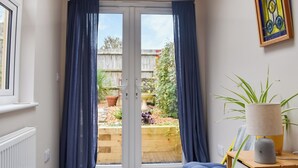 Une pièce dotée d’une porte-fenêtre coulissante donnant sur un jardin, d’une petite chaise jaune, d’une lampe et d’une table d’appoint en bois.