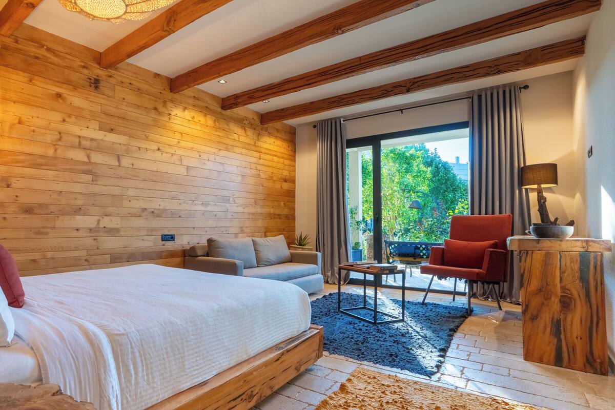 A bedroom with a wooden ceiling, a bed, a sofa, a red chair, a small table, and a view of greenery outside.
