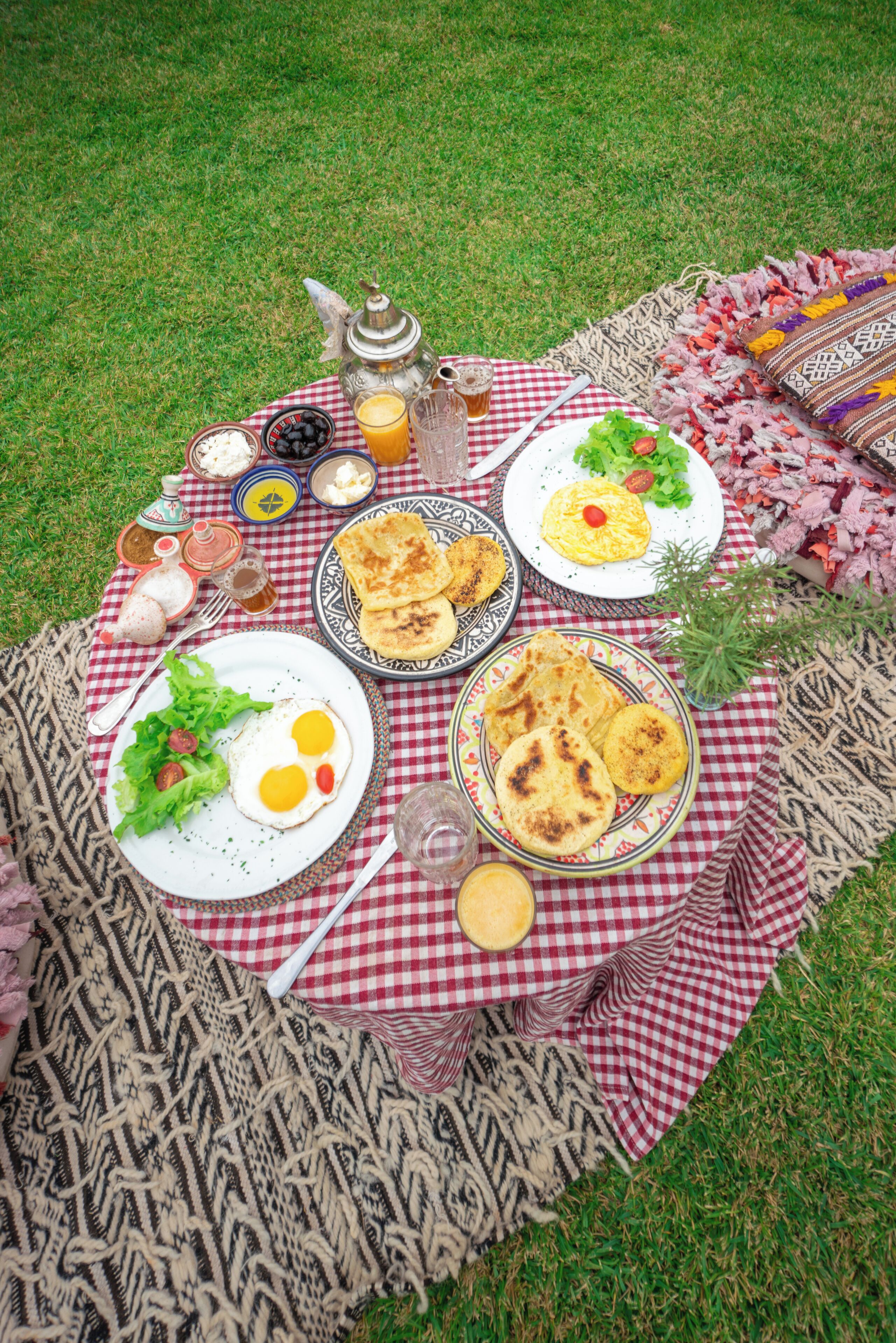 A picnic setup on a checkered blanket with plates of food, a teapot, and a glass of juice.