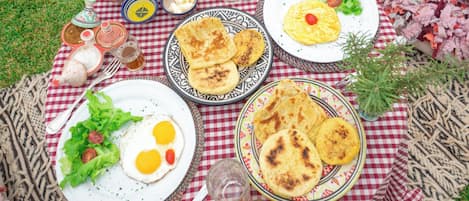 A picnic setup on a checkered blanket with plates of food, a teapot, and a glass of juice.