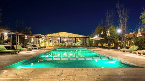 A nighttime swimming pool area with lounge chairs and umbrellas, surrounded by a wooden building with glass windows.