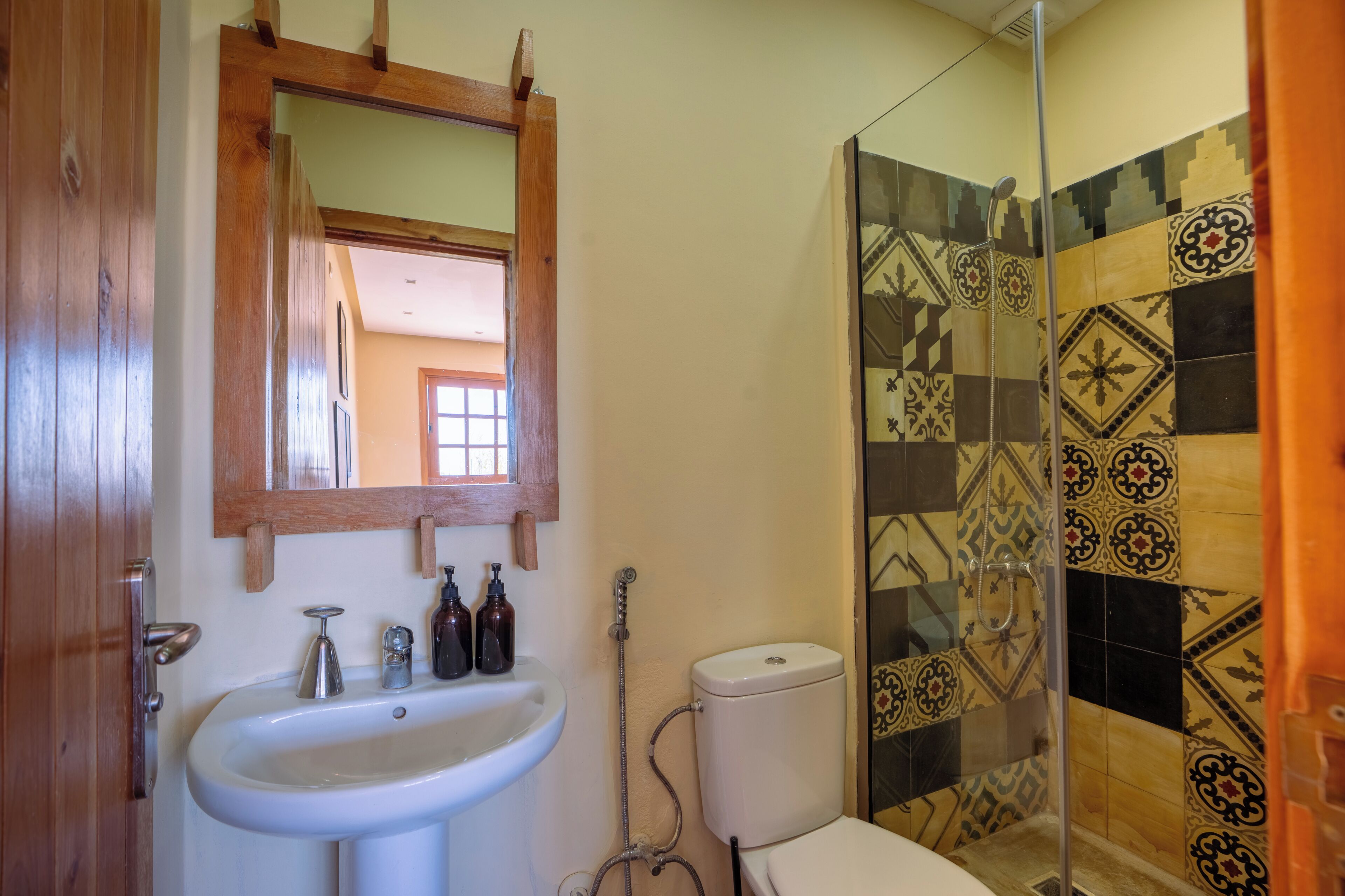 A bathroom with a wooden door, a white sink, a toilet, and a shower with patterned tiles.