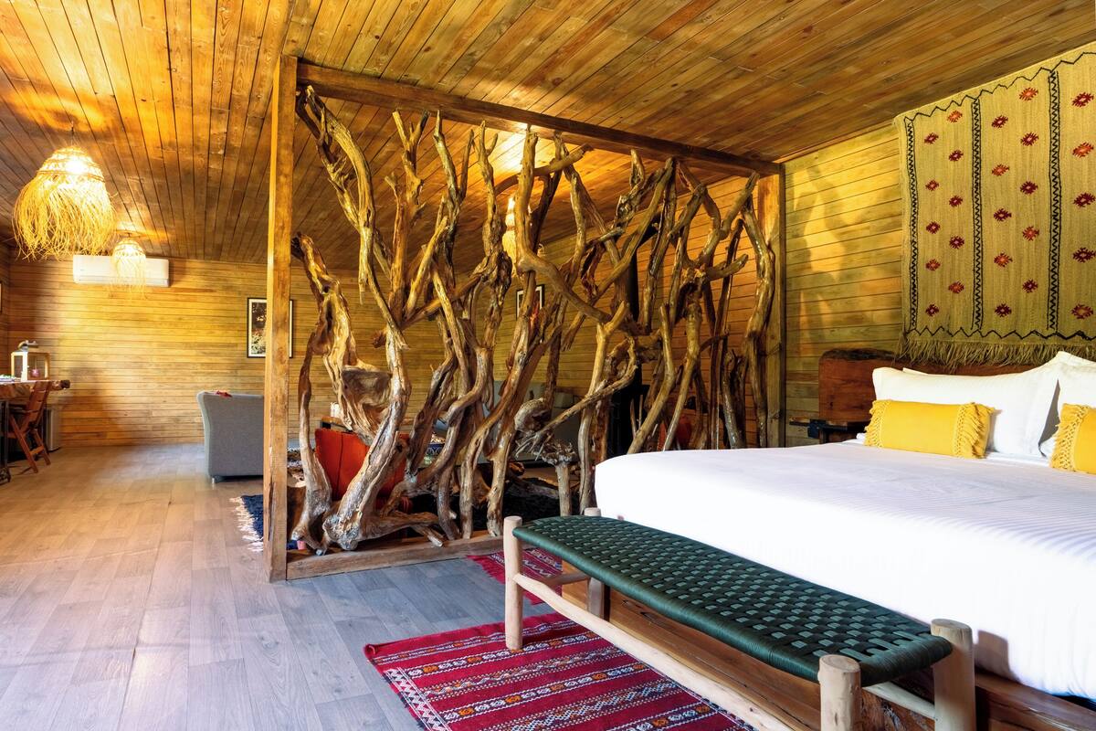 A wooden interior with a bed, a bench, a dining table, and a wall with a tree branch design.