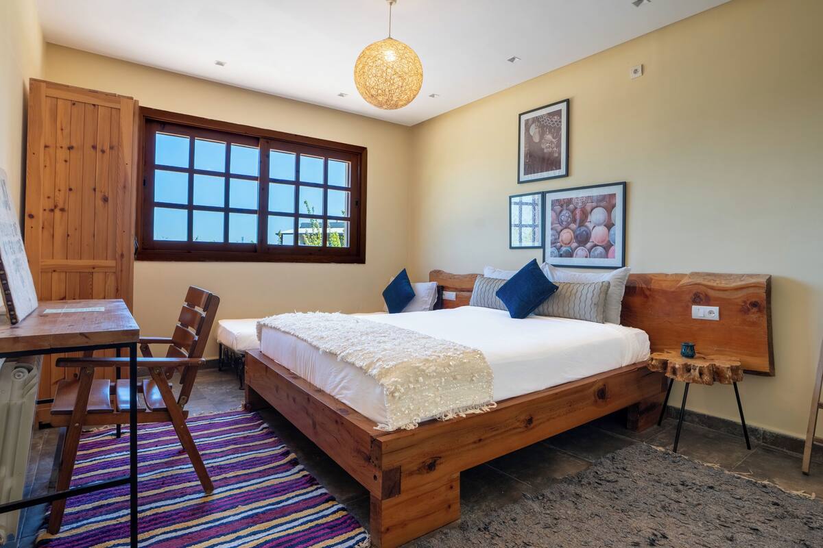 A bedroom with a wooden bed, a desk, a chair, a window, and a hanging light fixture.