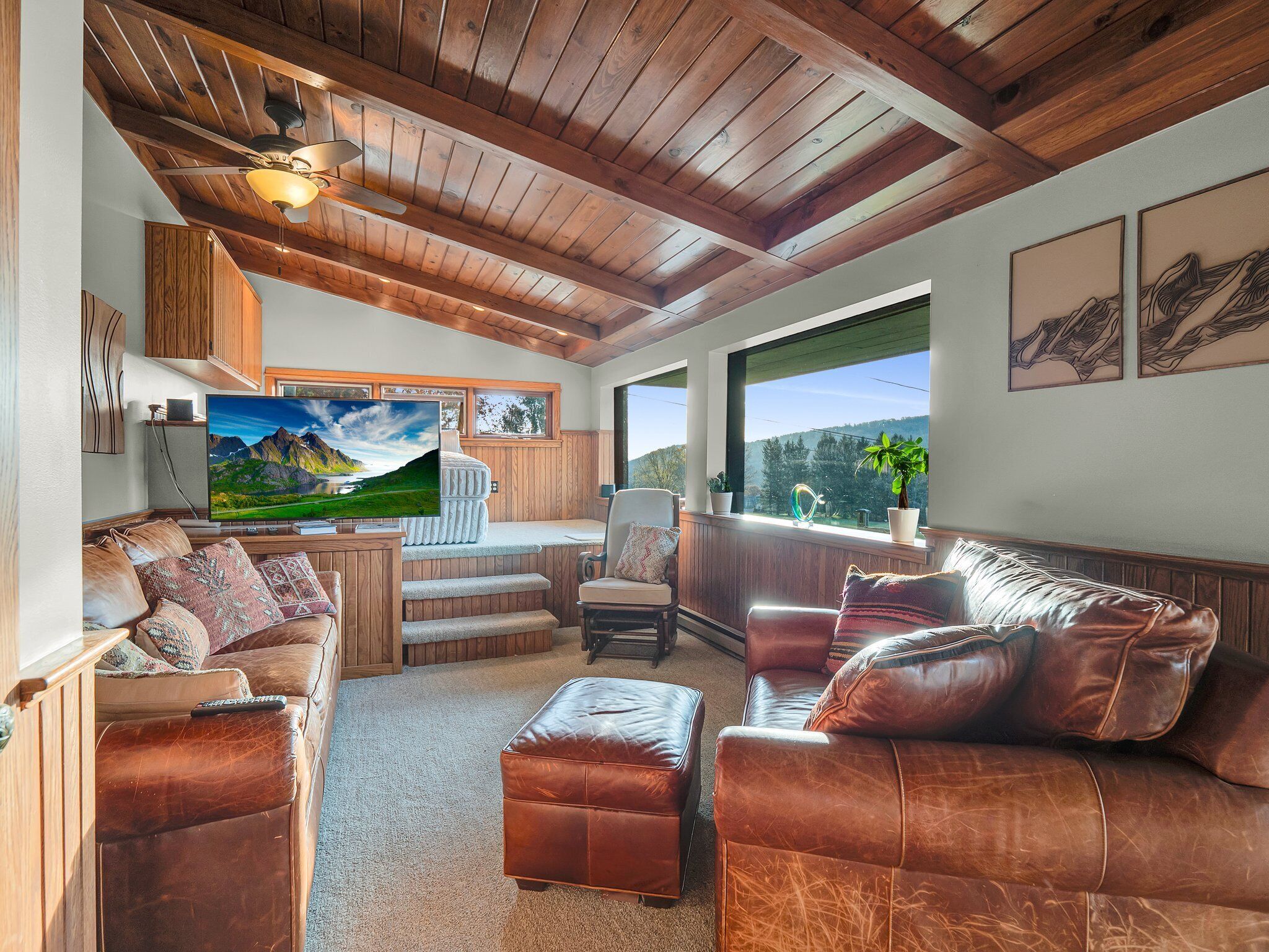 A cozy living room with a wooden ceiling, a large flat-screen TV, leather sofas, and a view of mountains through the windows.
