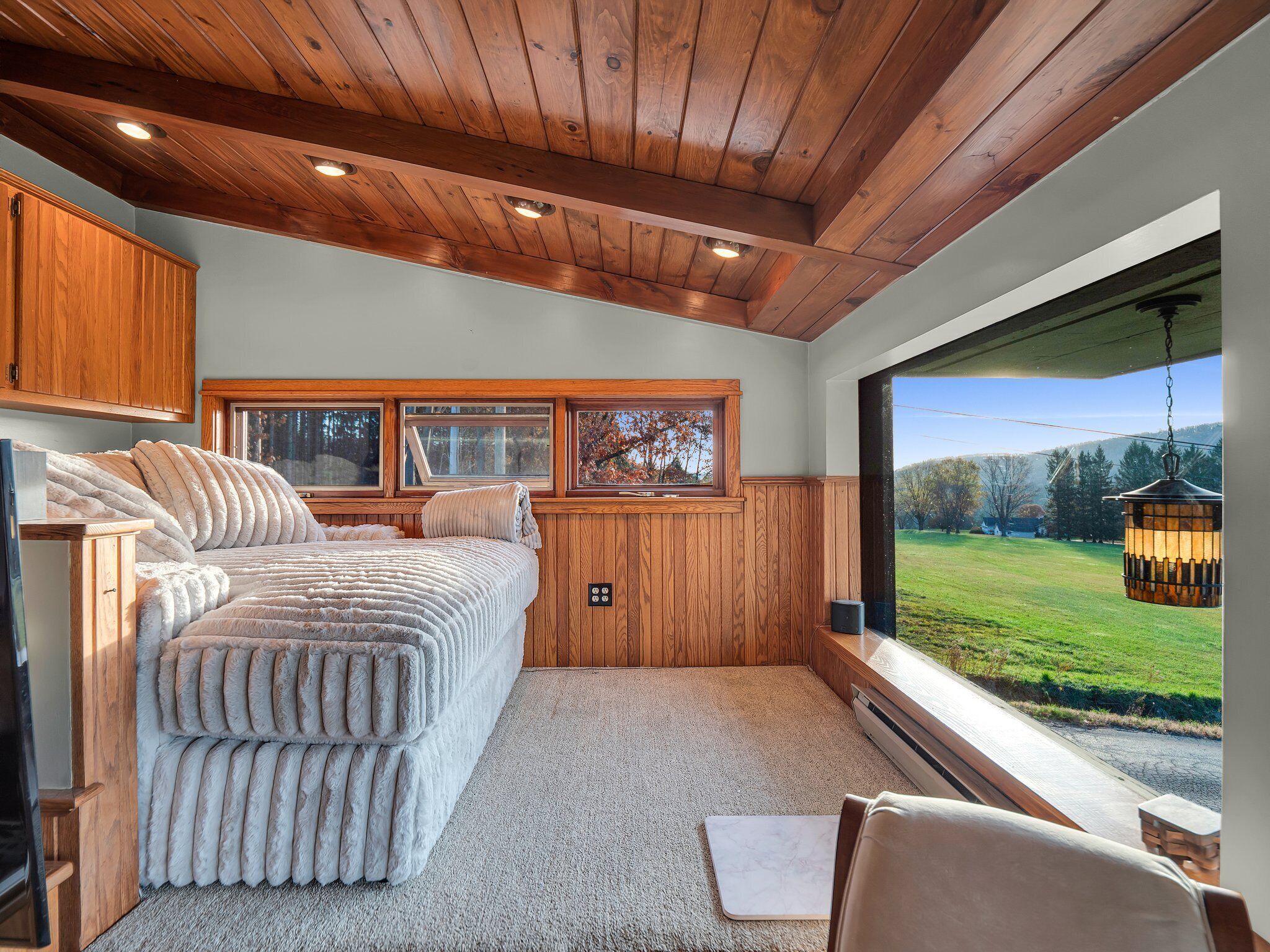 A bedroom with a large bed, wooden ceiling, and a view of a green landscape through a large window.