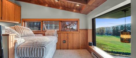 A bedroom with a large bed, wooden ceiling, and a view of a green landscape through a large window.