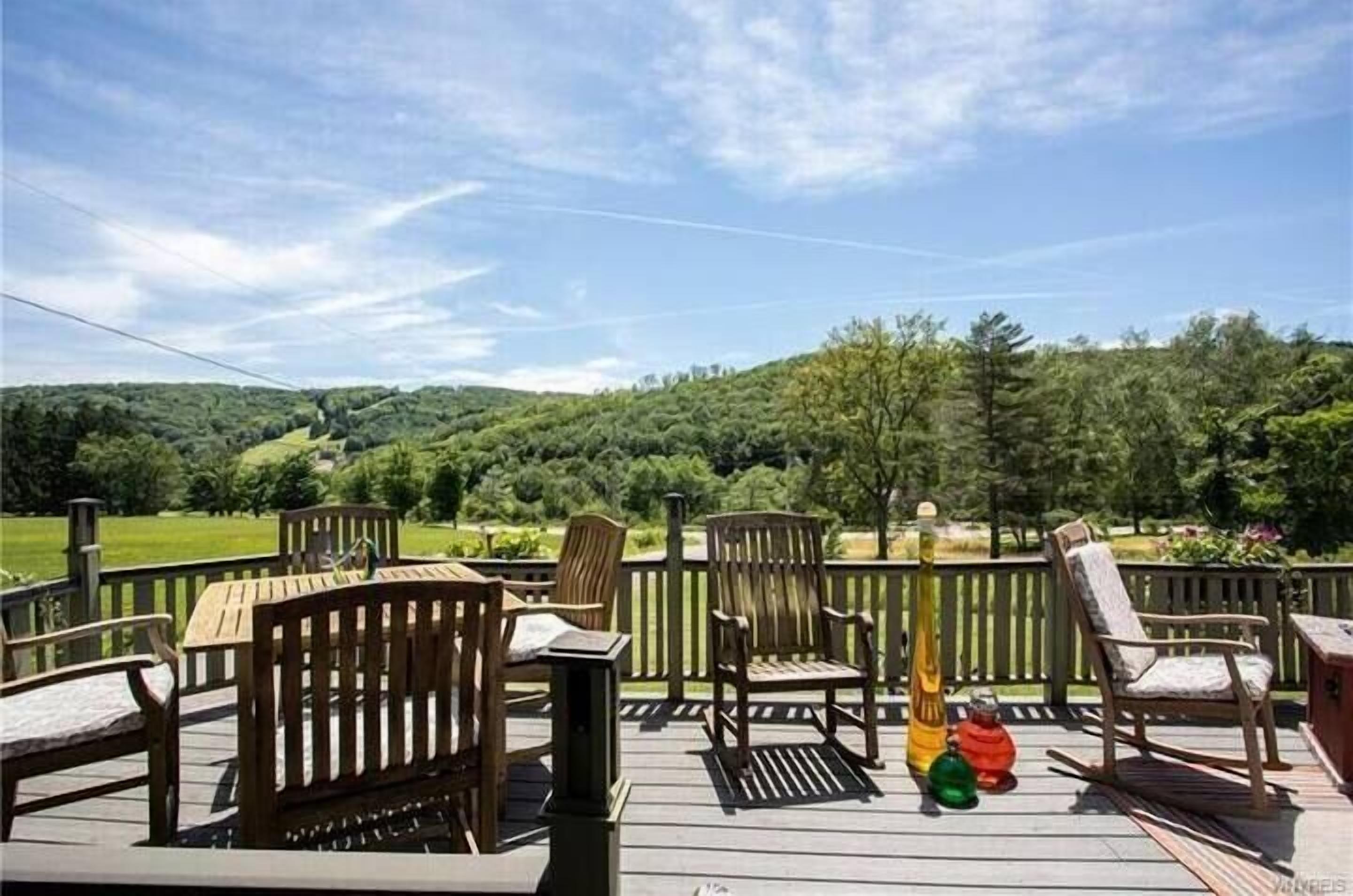 Een houten terras met buitenmeubilair, waaronder stoelen en een tafel, met uitzicht op een groen landschap met bomen en heuvels.