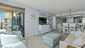 A modern living room with a sofa, ottoman, and a dining area with chairs and a table. - Windemere 605 -Oceanfront 3BR/2BA-Indoor Pool (North Myrtle Beach)