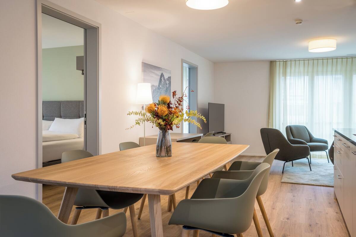 A modern hotel room with a wooden dining table, green chairs, a vase with flowers, a flat-screen TV, and a view of the outdoors through large windows.