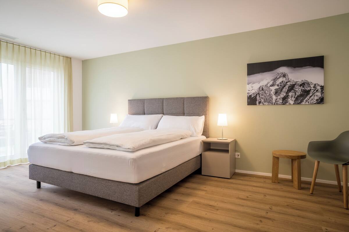 A neatly made bed with a grey headboard, a bedside table with a lamp, a wooden stool, and a chair.