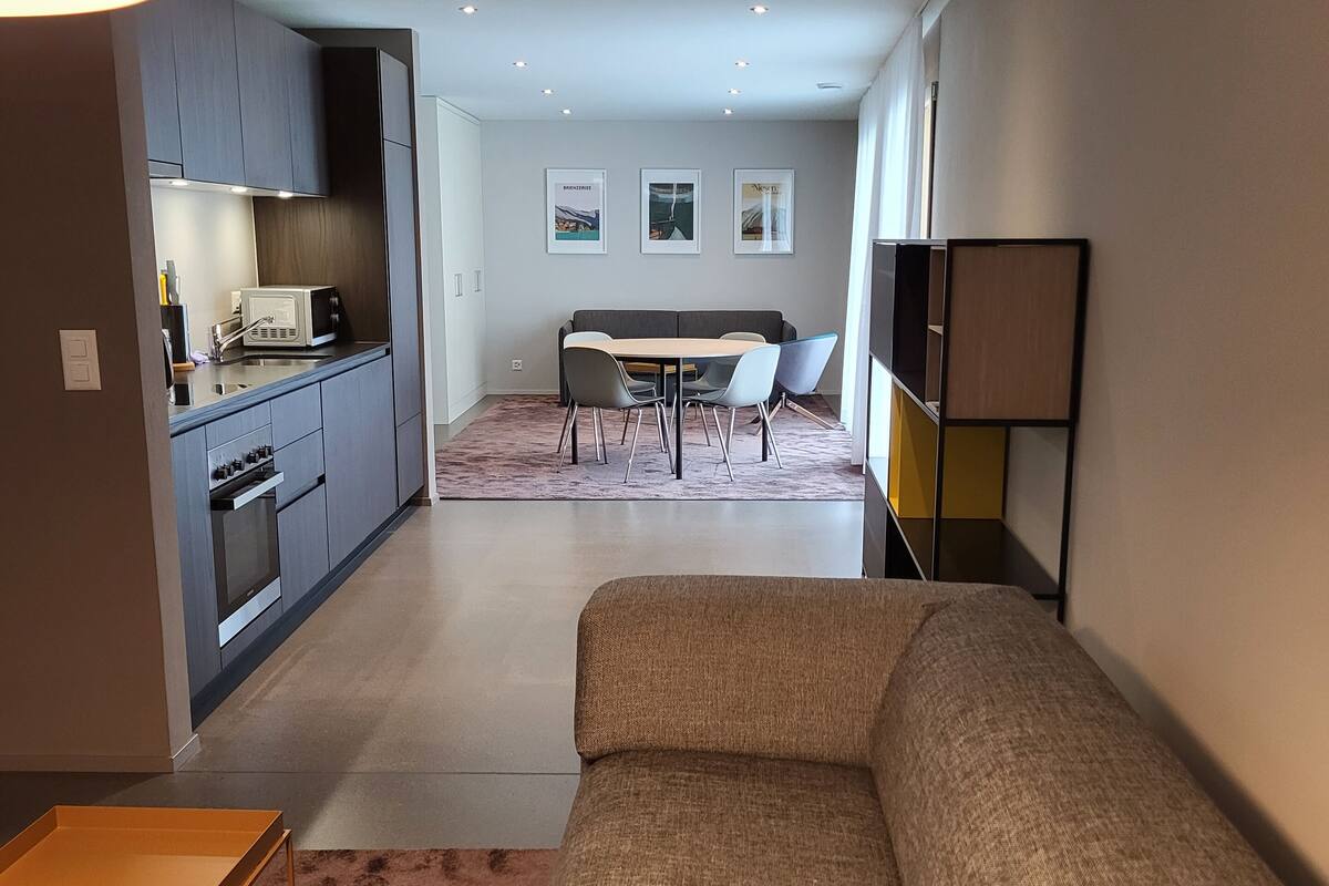 A modern living room with a beige sofa, a small table, and a kitchen area with cabinets and a microwave. There is a dining area with a table and chairs in the background.