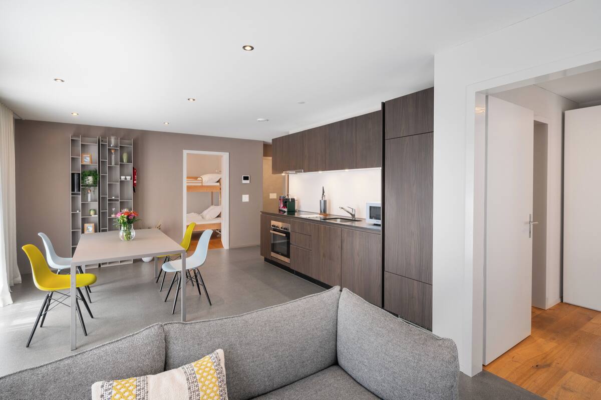 A modern living room with a grey sofa, a dining area with a table and chairs, and a kitchen with dark wood cabinets and a white countertop.