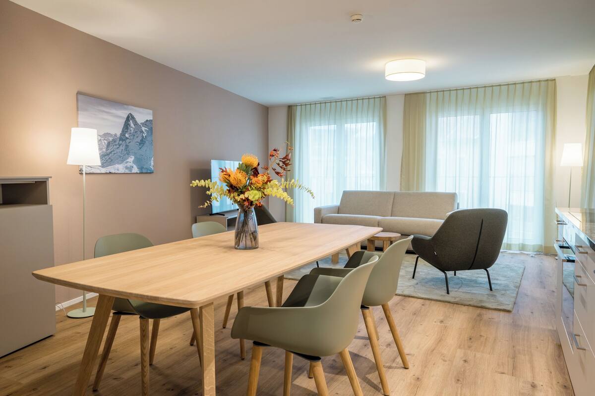 A modern living room with a wooden dining table, green chairs, a sofa, and a TV.