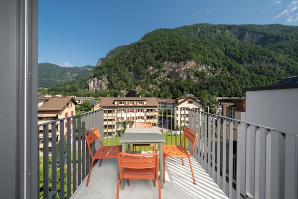 A balcony with a table and chairs, overlooking a residential area with houses and a forested mountain.