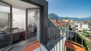A balcony with a glass door leading to a room with a sofa and a table. The balcony overlooks a residential area with houses and mountains in the background. - Modern City Apartments (Interlaken)