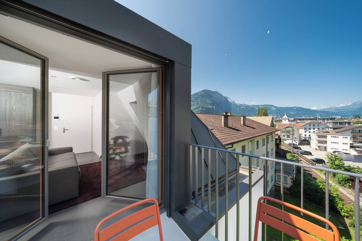A balcony with a glass door leading to a room with a sofa and a table. The balcony overlooks a residential area with houses and mountains in the background.