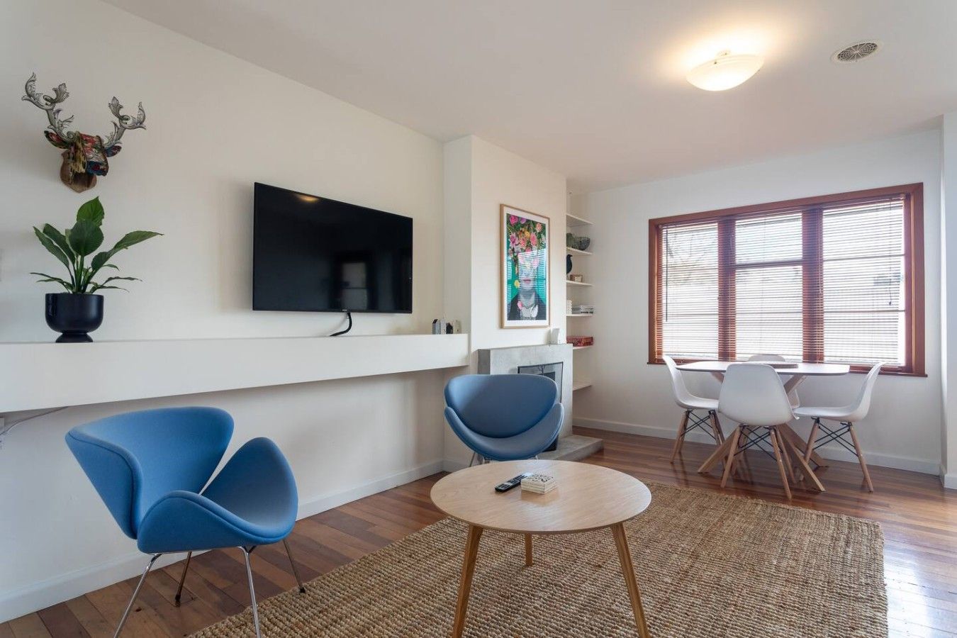 A modern living room with a flat-screen TV, blue chairs, a round coffee table, and a large window with blinds.