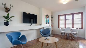 A modern living room with a flat-screen TV, blue chairs, a round coffee table, and a large window with blinds.