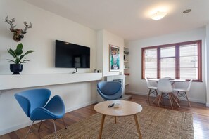 A modern living room with a flat-screen TV, blue chairs, a round coffee table, and a large window with blinds.