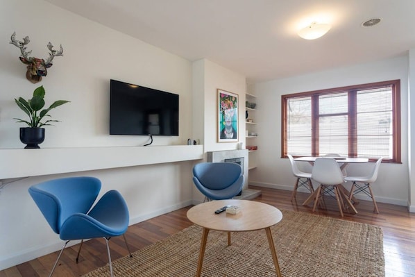 A modern living room with a flat-screen TV, blue chairs, a round coffee table, and a large window with blinds.