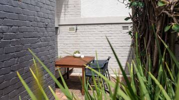 A small outdoor seating area with a table and chairs, surrounded by a brick wall and vegetation.