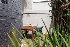 A small outdoor seating area with a table and chairs, surrounded by a brick wall and vegetation.