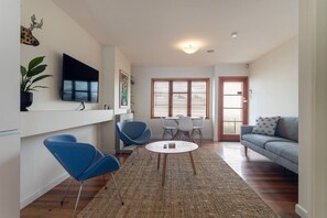 A modern living room with a flat-screen TV, blue chairs, a gray sofa, and a large window with blinds.