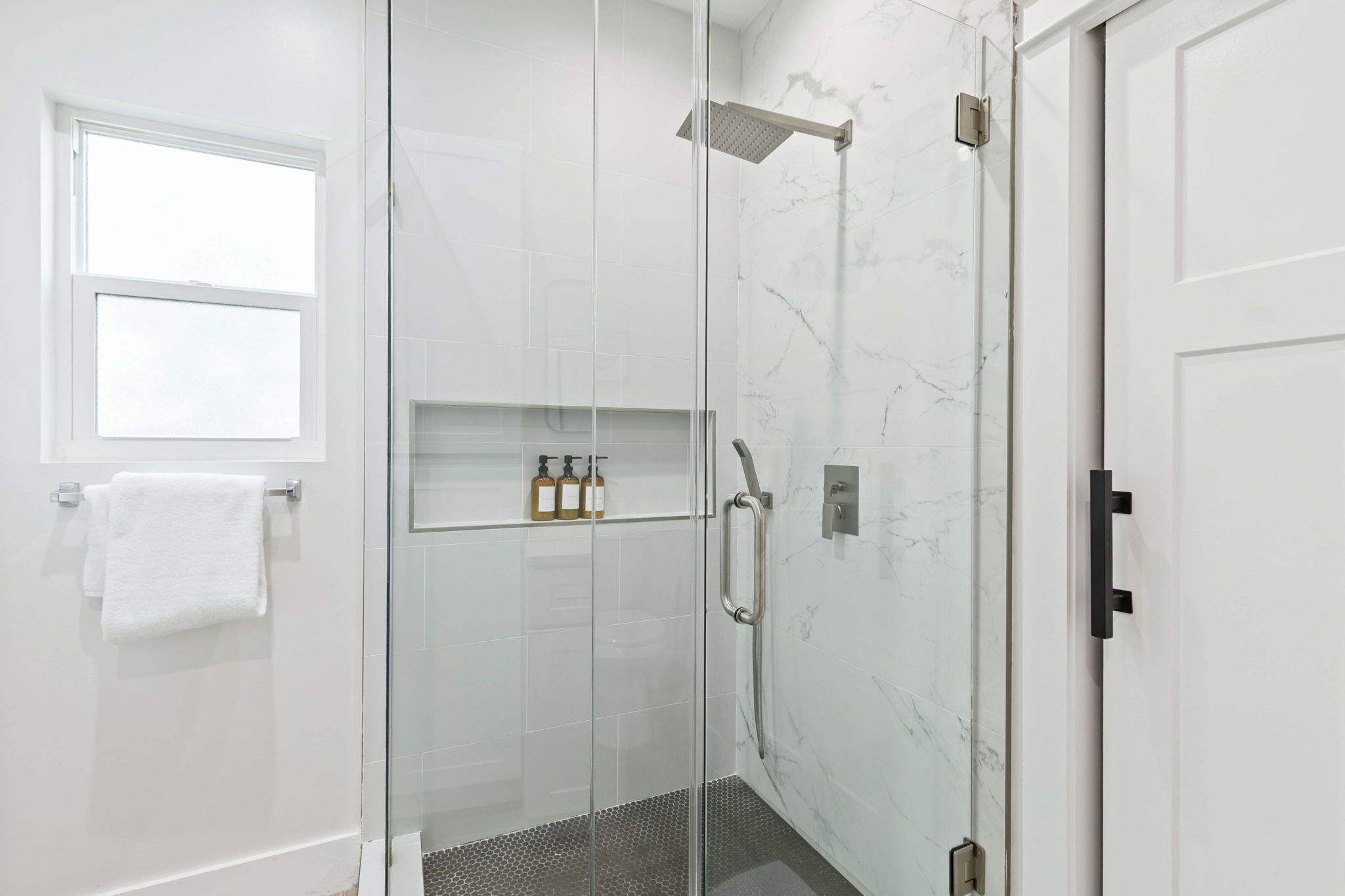 Un baño moderno con ducha de vidrio, pared de azulejos blancos, un estante pequeño con artículos de aseo y una ventana sobre el perchero.