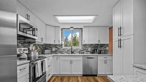 Une cuisine moderne avec des meubles blancs, un lave-vaisselle, une cuisinière et une fenêtre donnant sur les arbres.