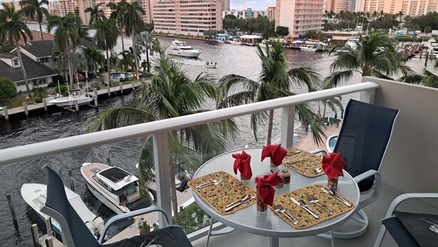 A balcony with a table set for dining, overlooking a marina with boats and palm trees, and high-rise buildings in the background.