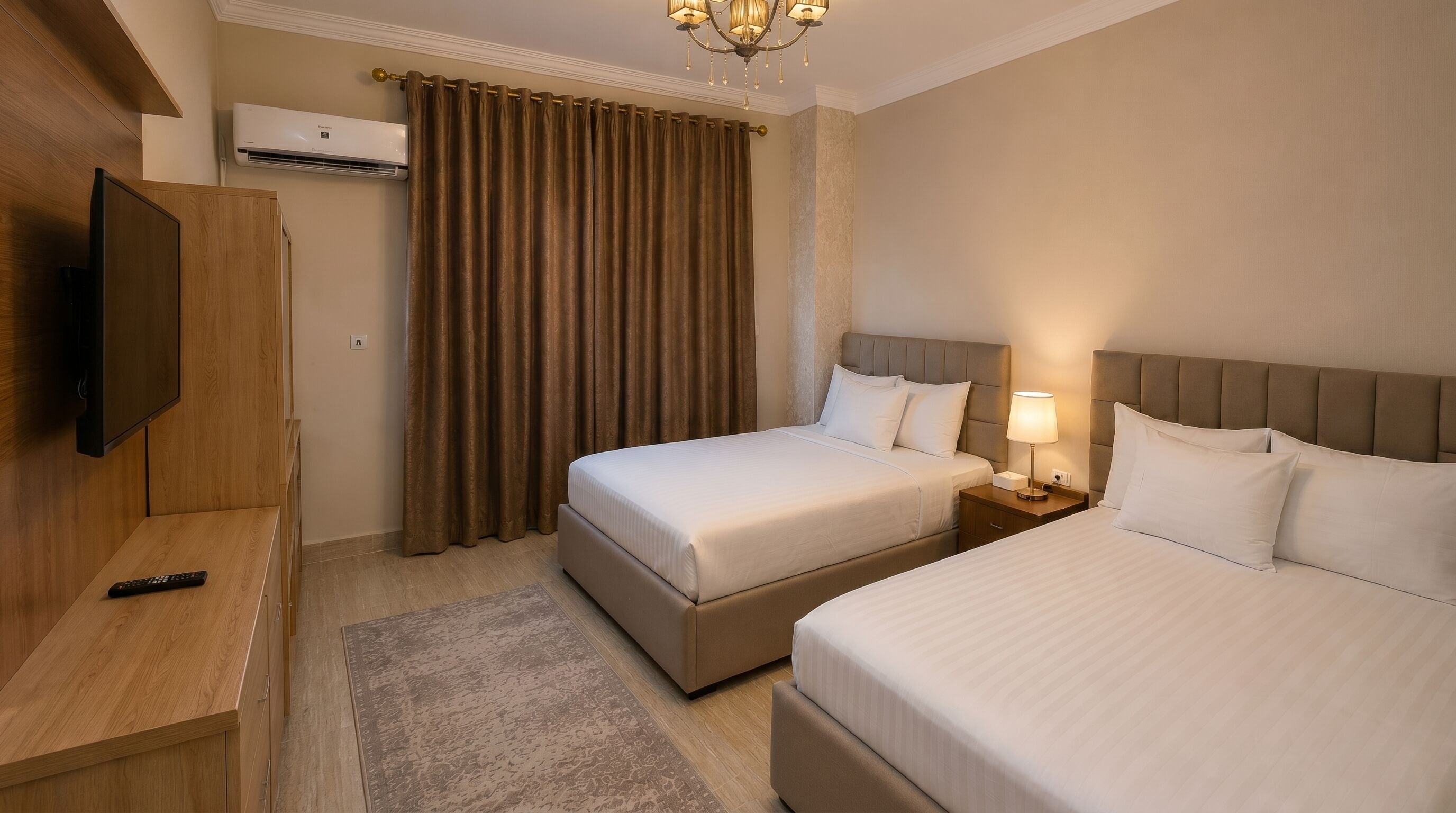 A hotel room with two beds, a TV, a bedside table, a lamp, and a window with curtains.