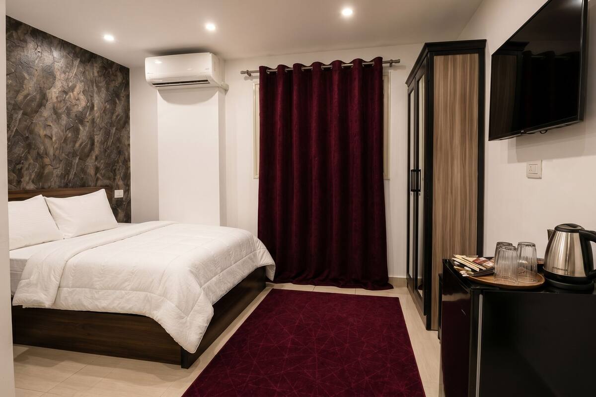 A hotel room with a bed, a red curtain, a TV, a coffee maker, and a small table with glasses and books.