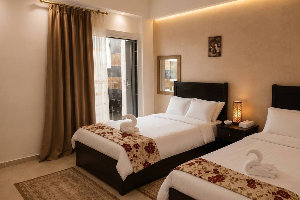 A hotel room with two beds, a nightstand with a lamp, a window with curtains, and a wall-mounted picture.