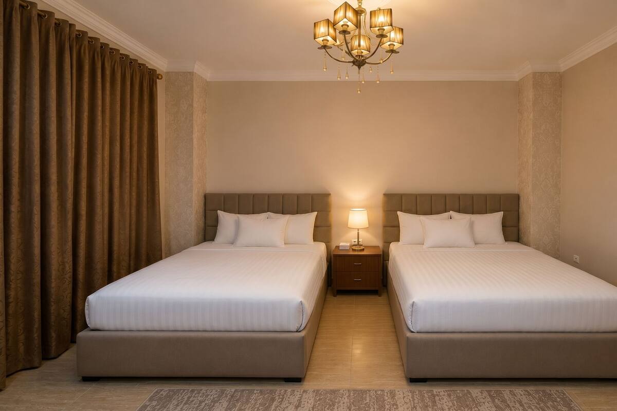 A hotel room with two beds, a chandelier, a nightstand with a lamp, and a patterned curtain.
