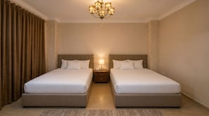 A hotel room with two beds, a chandelier, a nightstand with a lamp, and a patterned curtain.