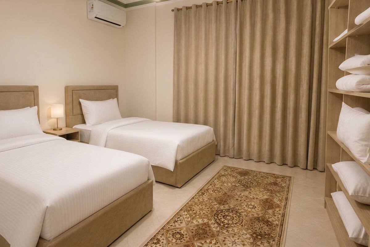 A hotel room with two beds, a nightstand, a lamp, a large window with curtains, and a built-in shelf with pillows.
