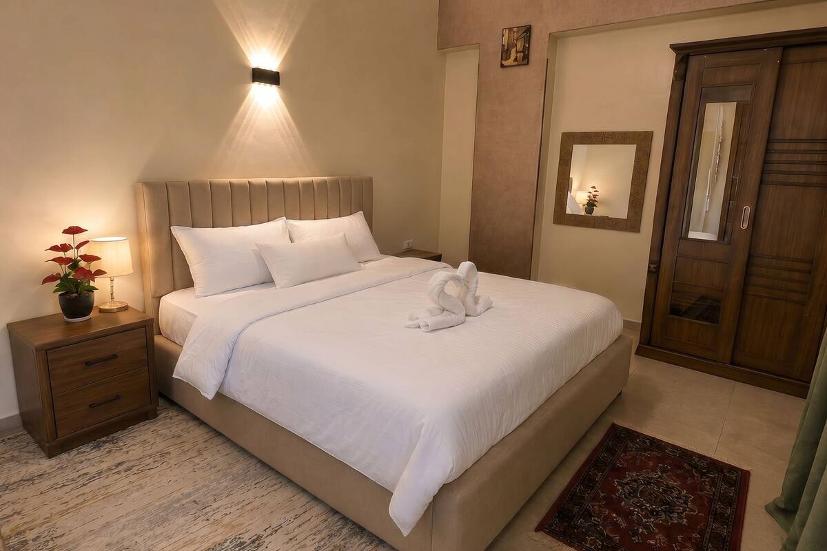 A neatly made bed with white linens, a bedside lamp, a potted plant, and a wooden nightstand.
