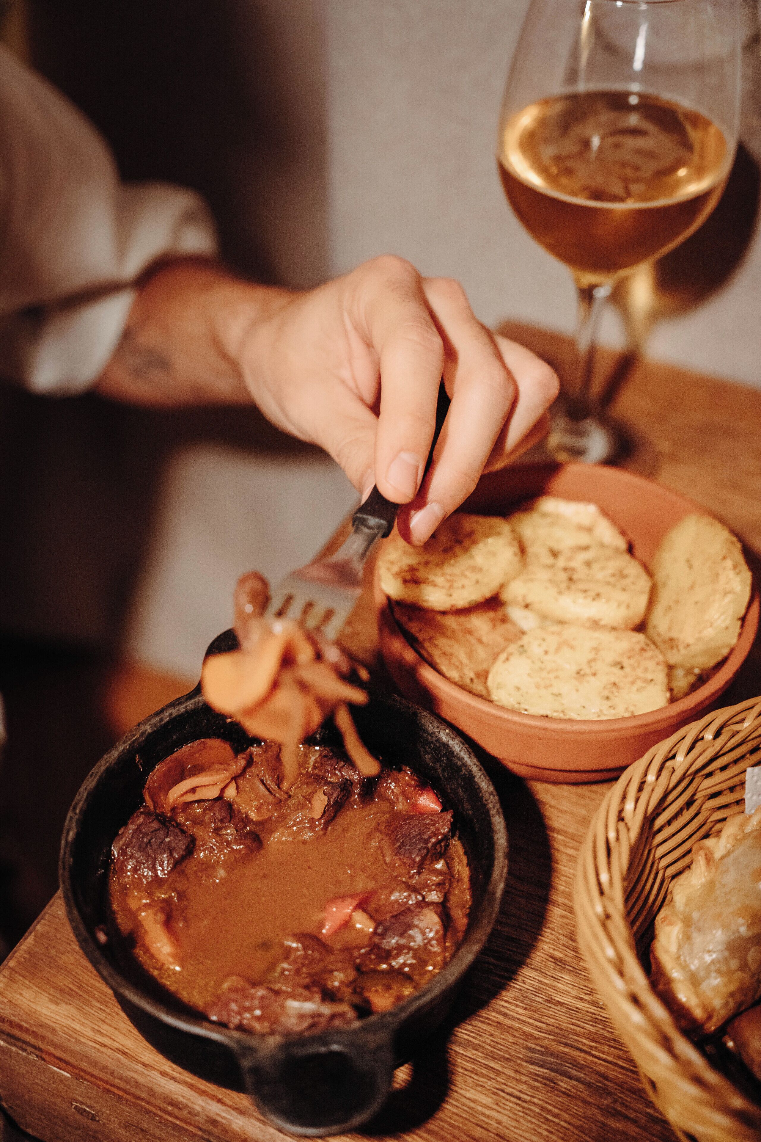 Una persona sirviendo comida desde una sartén de hierro fundido, un bol de bizcochos y una copa de vino sobre una mesa de madera.