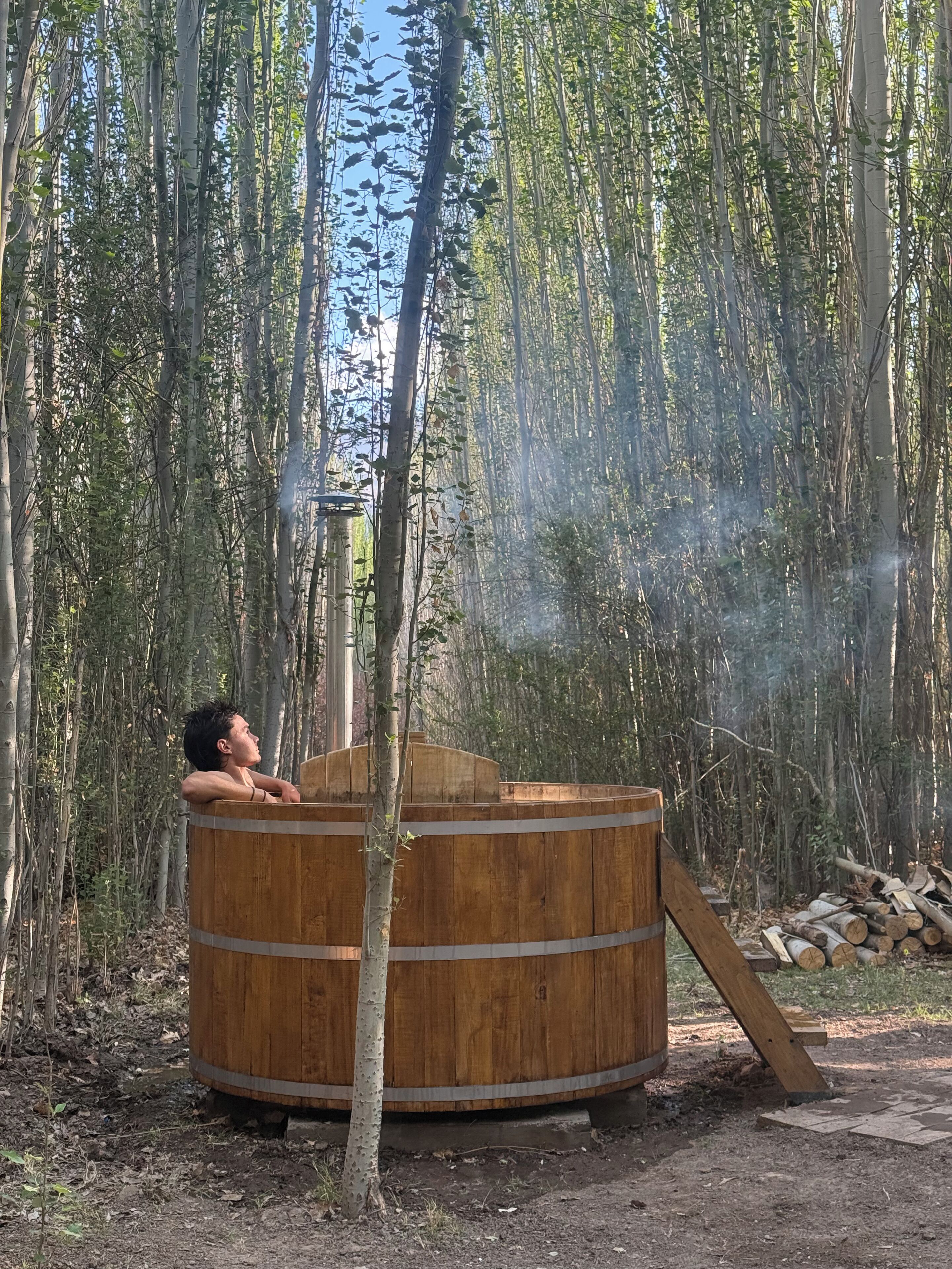 A wooden hot tub in a forested area with a person relaxing inside.