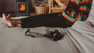 A person sitting on a bed in a room with a wood-burning stove, a shelf with a lamp, and a camera on the bed.