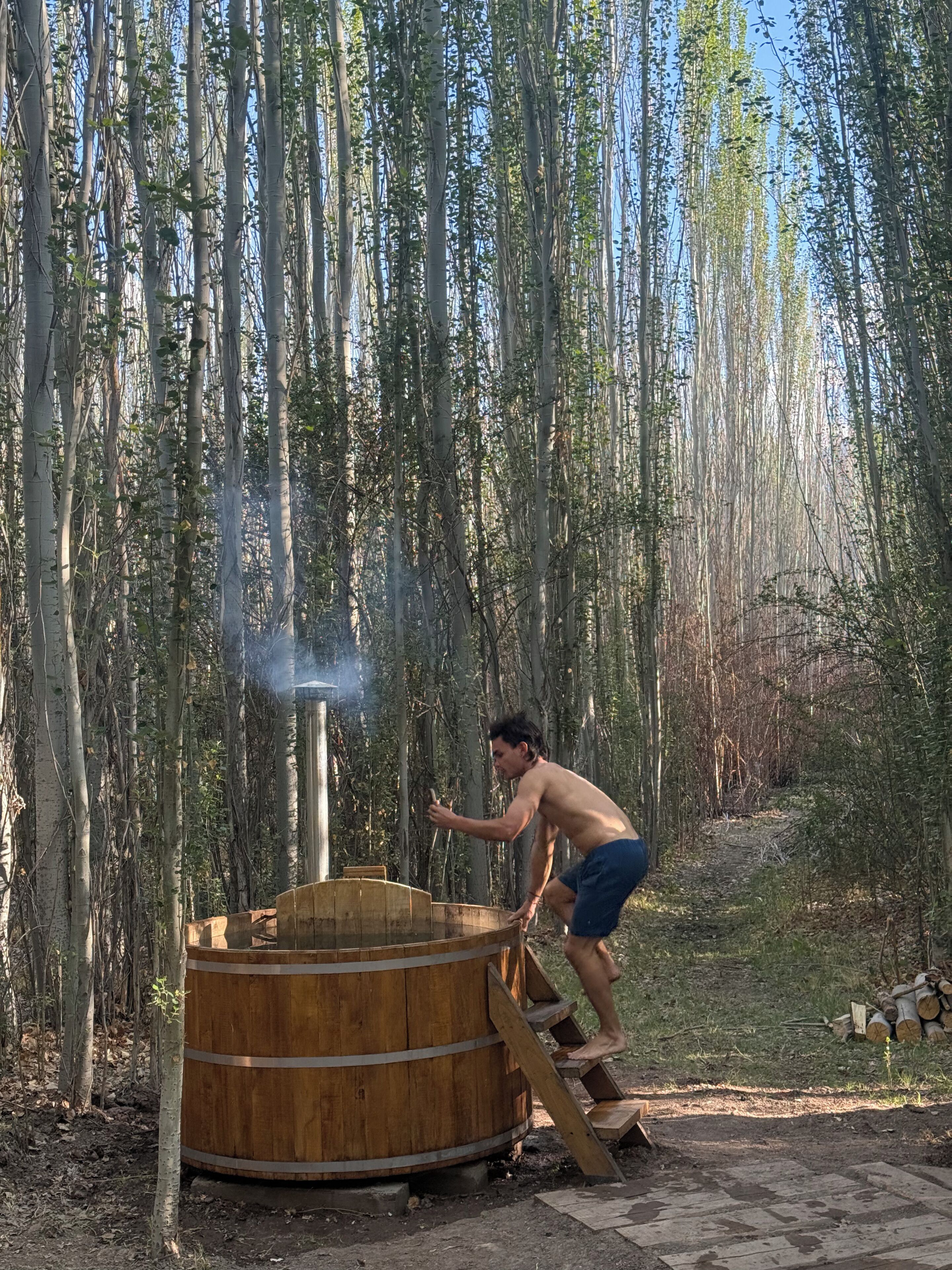 A person using a sauna in a forested area with a wooden structure and a chimney emitting steam.