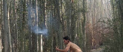 A person using a sauna in a forested area with a wooden structure and a chimney emitting steam.