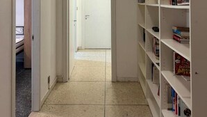 A narrow hallway with a tiled floor leading to a door. A white bookshelf is on the right side.